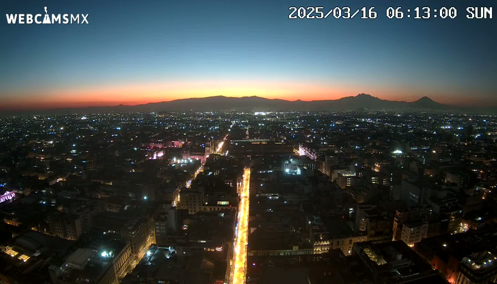 A nighttime aerial view of a city with brightly lit streets and buildings, showing a sunset with mountains in the background.