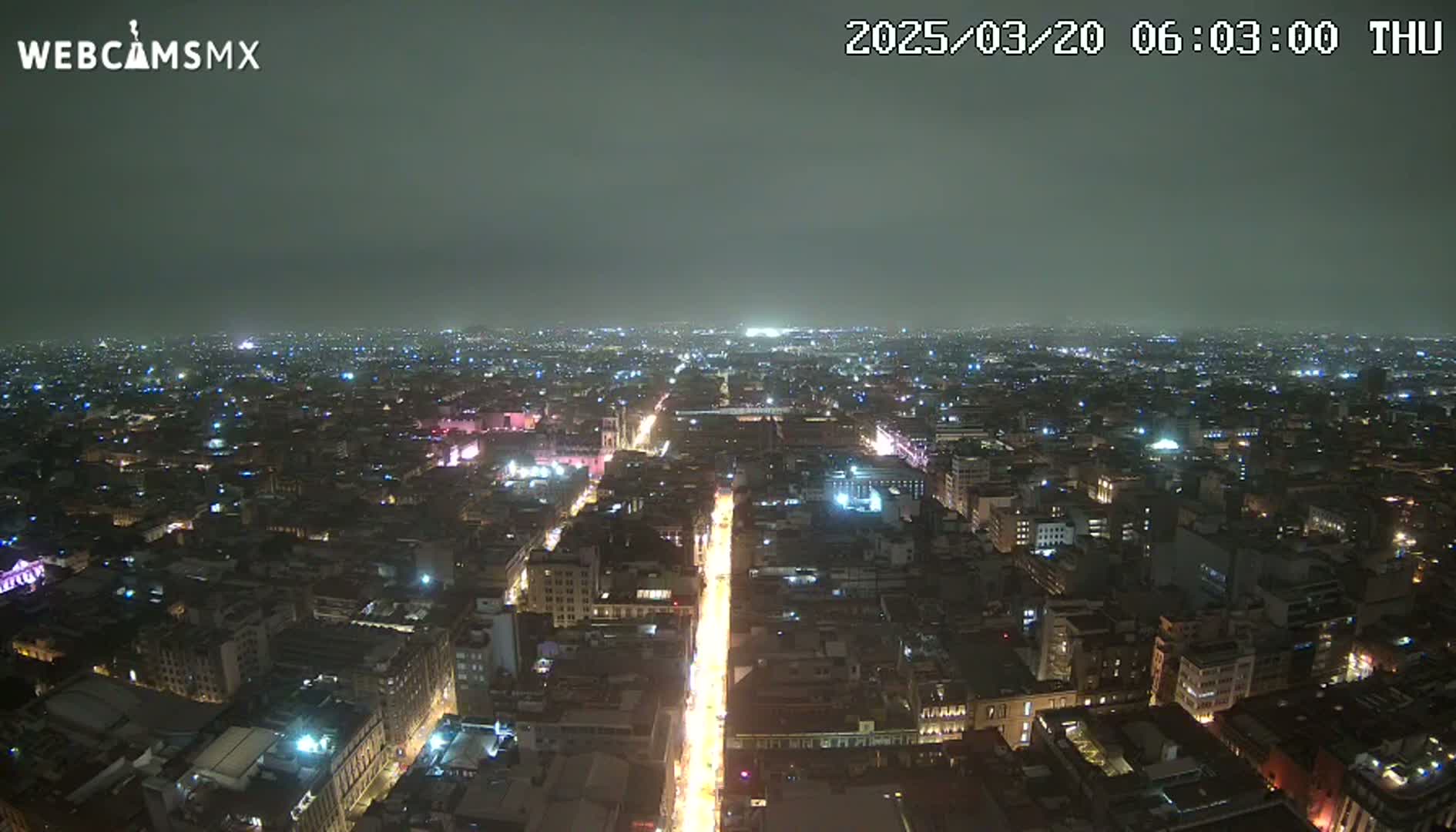 A nighttime aerial view of a city under hazy or cloudy conditions shows a long, brightly lit street stretching into the distance, surrounded by numerous buildings with visible lights.