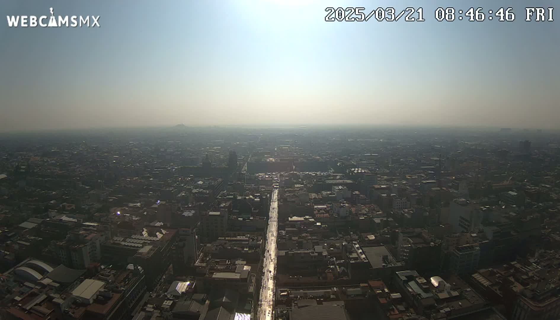 A hazy, sunny aerial view of a sprawling city, showing a long street extending from the foreground into the distance.