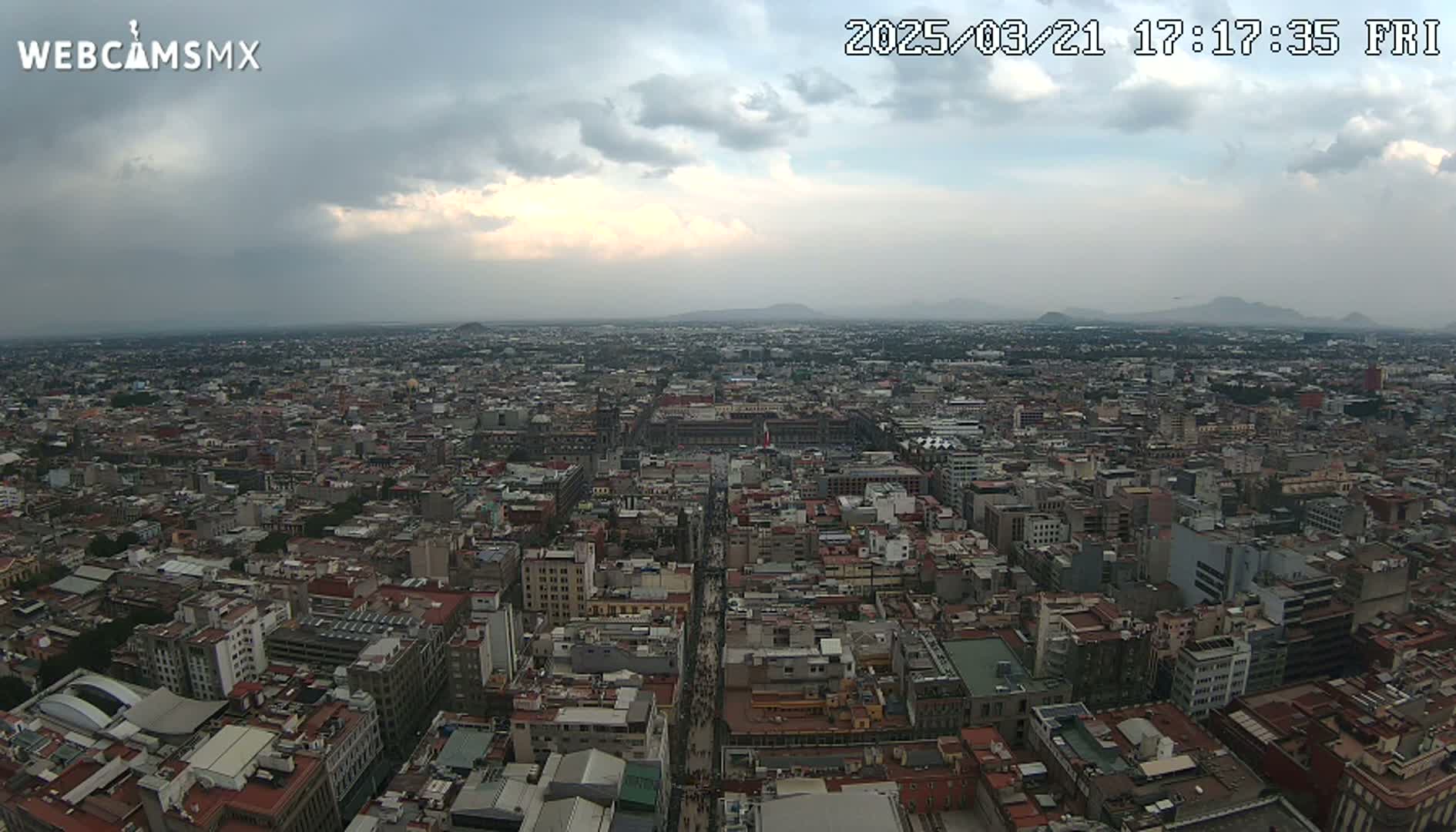 A high-angle view of a sprawling city under a cloudy sky shows a dense collection of buildings extending to distant mountains.