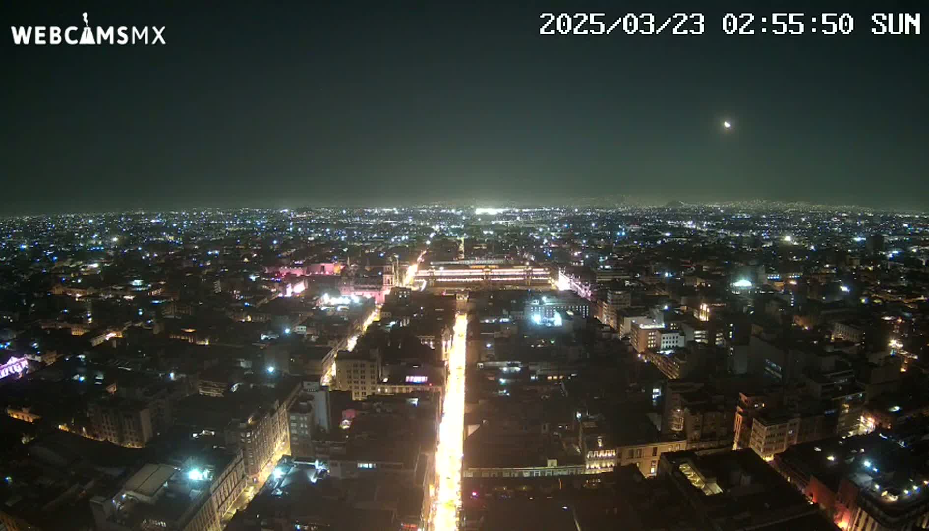 A nighttime, high-angle view shows a large city with brightly lit streets and buildings under a clear sky with a visible moon.
