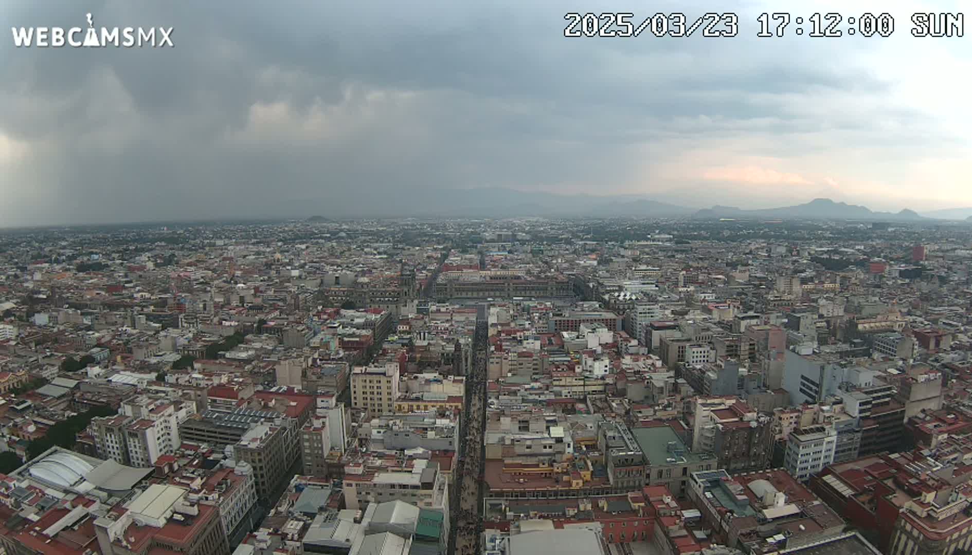 A high-angle view of a sprawling city under a mostly cloudy sky, with mountains visible in the distance.