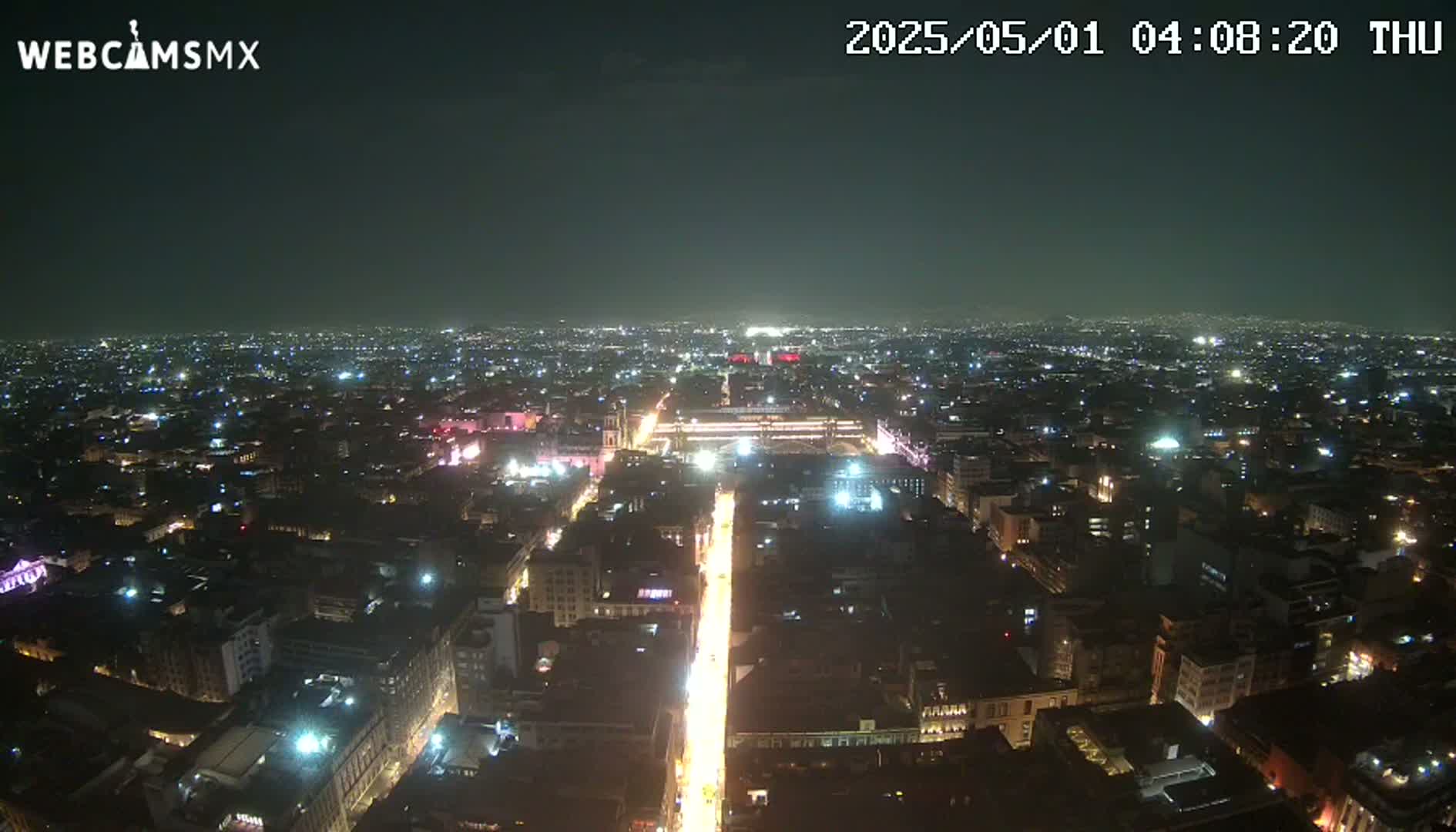 A brightly lit, wide city street at night is seen from above, surrounded by a sprawling cityscape under a clear, dark sky.