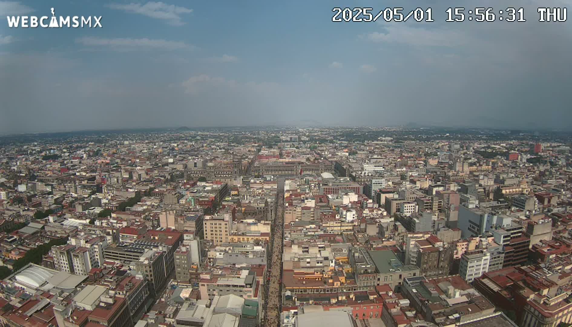 A high-angle, long shot on a partly cloudy day shows a sprawling cityscape of densely packed buildings extending to the horizon.