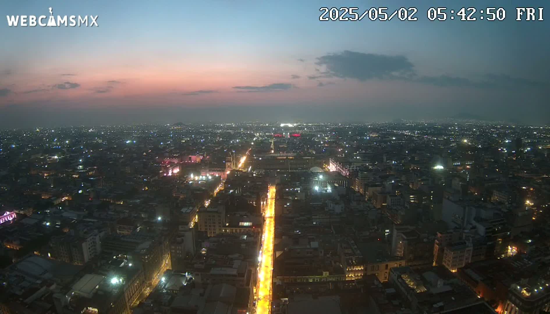 A brightly lit city street at twilight is visible from an elevated perspective, showing the surrounding cityscape under a mostly clear sky.