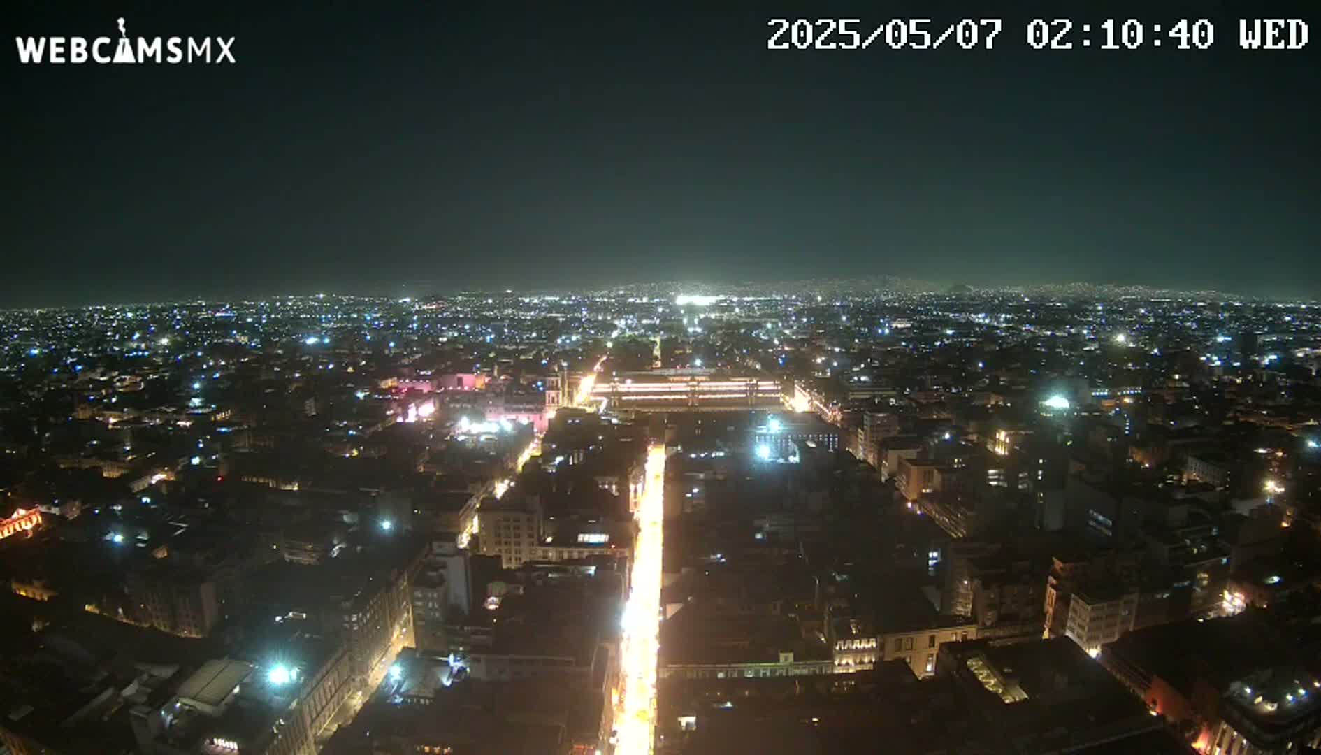 A brightly lit city street at night stretches toward a sprawling, dimly lit cityscape under a clear, dark sky.