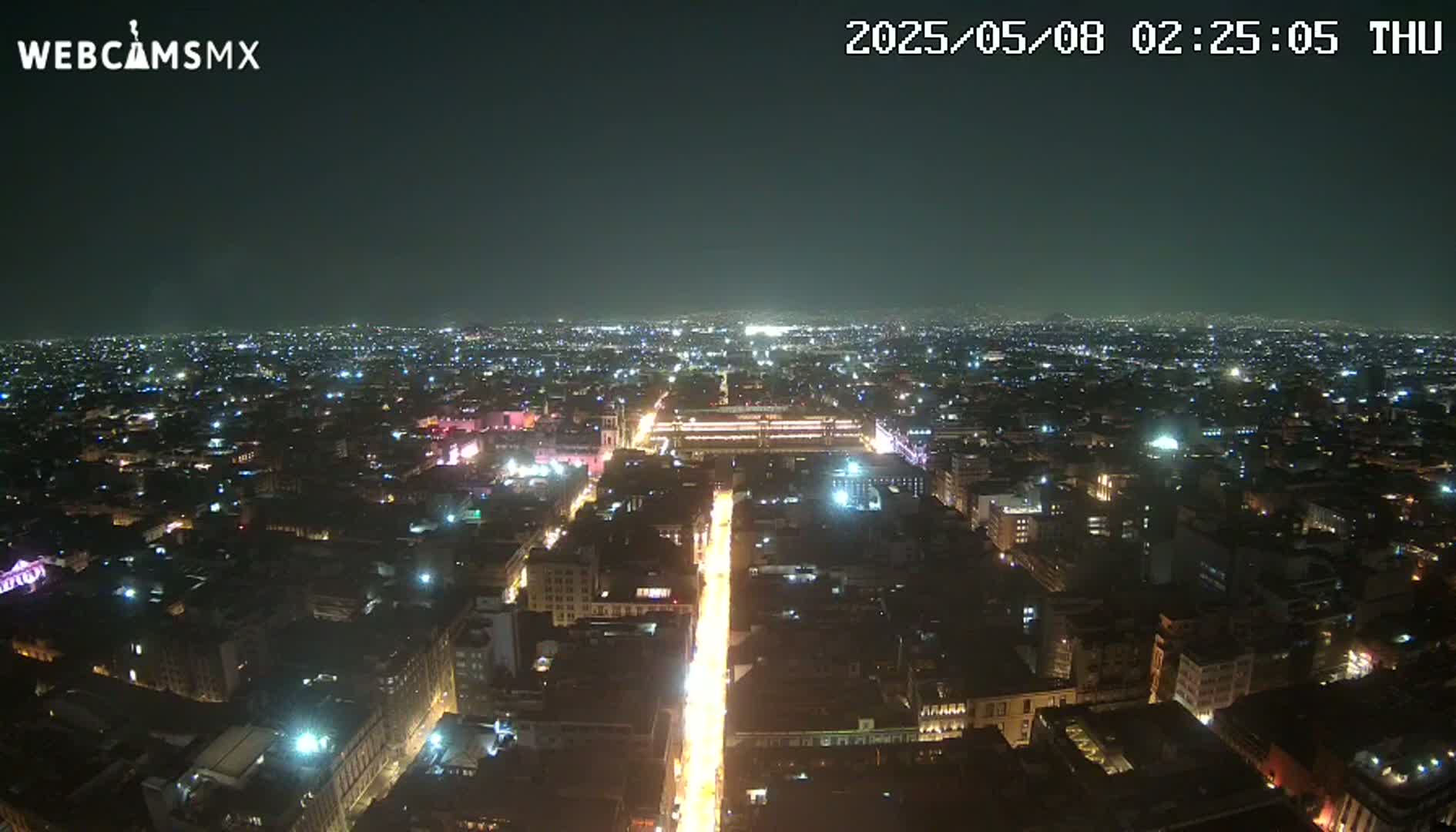 A brightly lit city street at night extends from a wide, illuminated plaza toward a sprawling, dark cityscape under a clear sky.