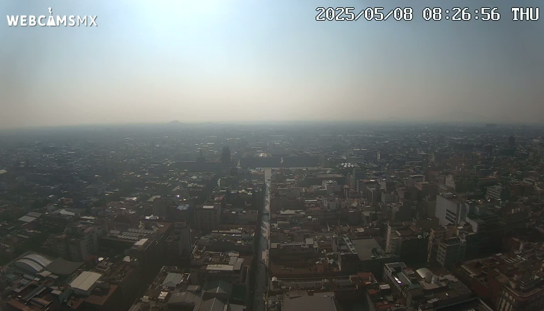 A hazy, sunny aerial view shows a sprawling city with numerous buildings and streets.