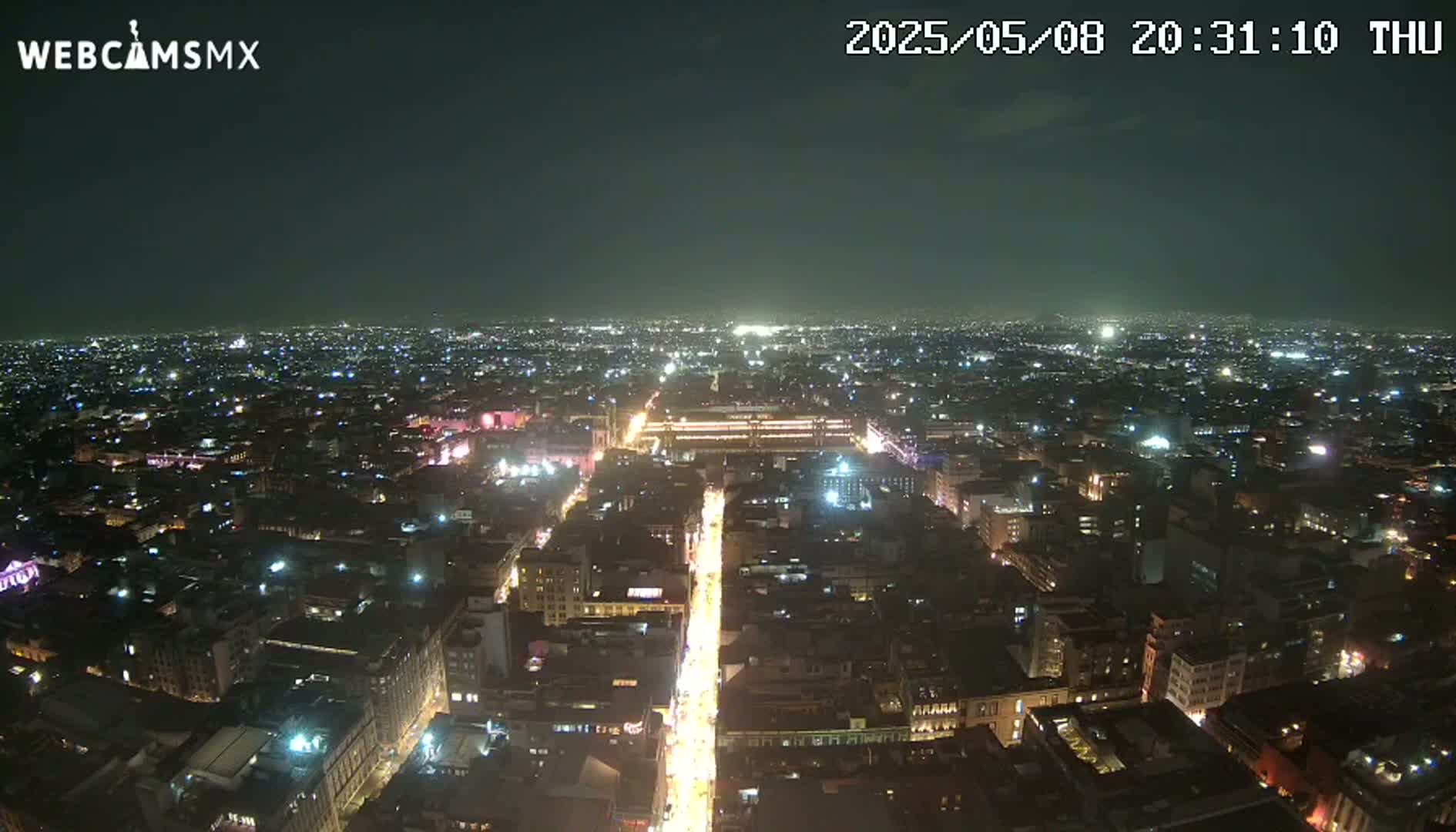 A nighttime, high-angle view shows a sprawling city illuminated by countless lights, with a brightly lit avenue cutting through the urban landscape.