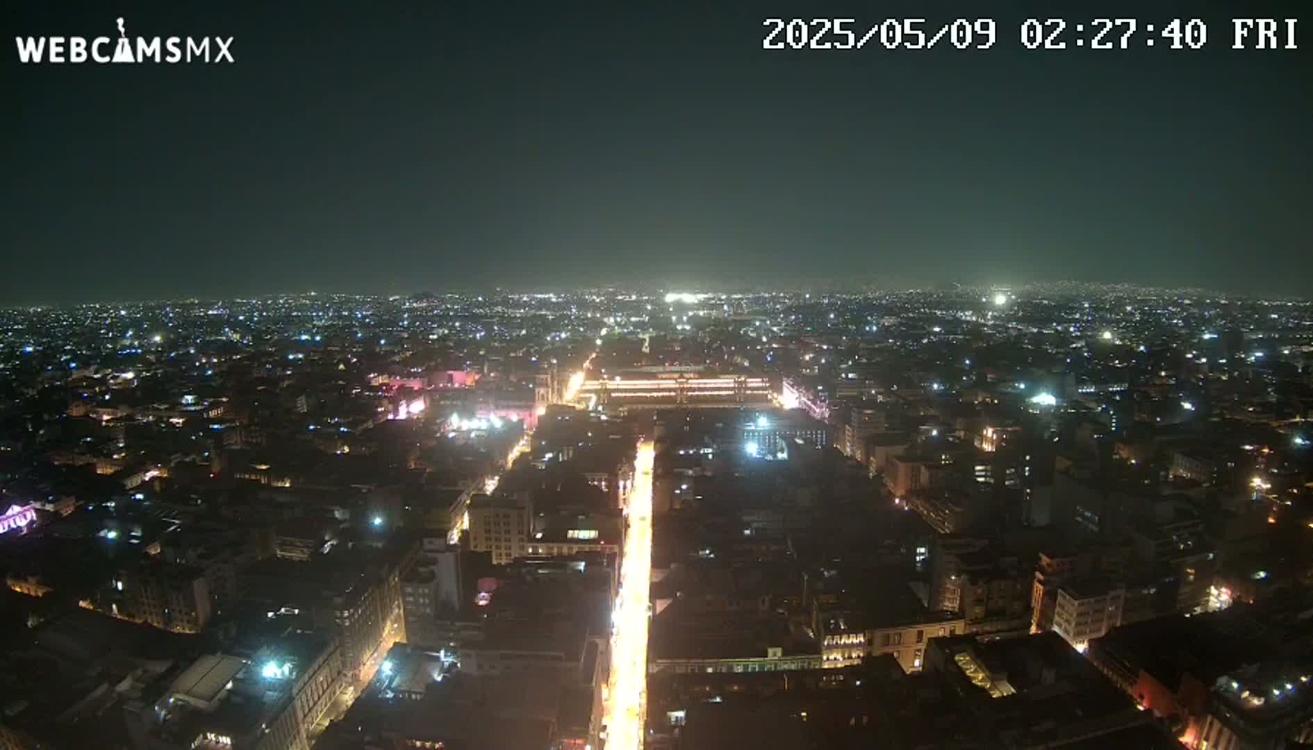 A brightly lit city street at night stretches out from a high vantage point, surrounded by a vast expanse of dimly lit buildings under a clear, dark sky.