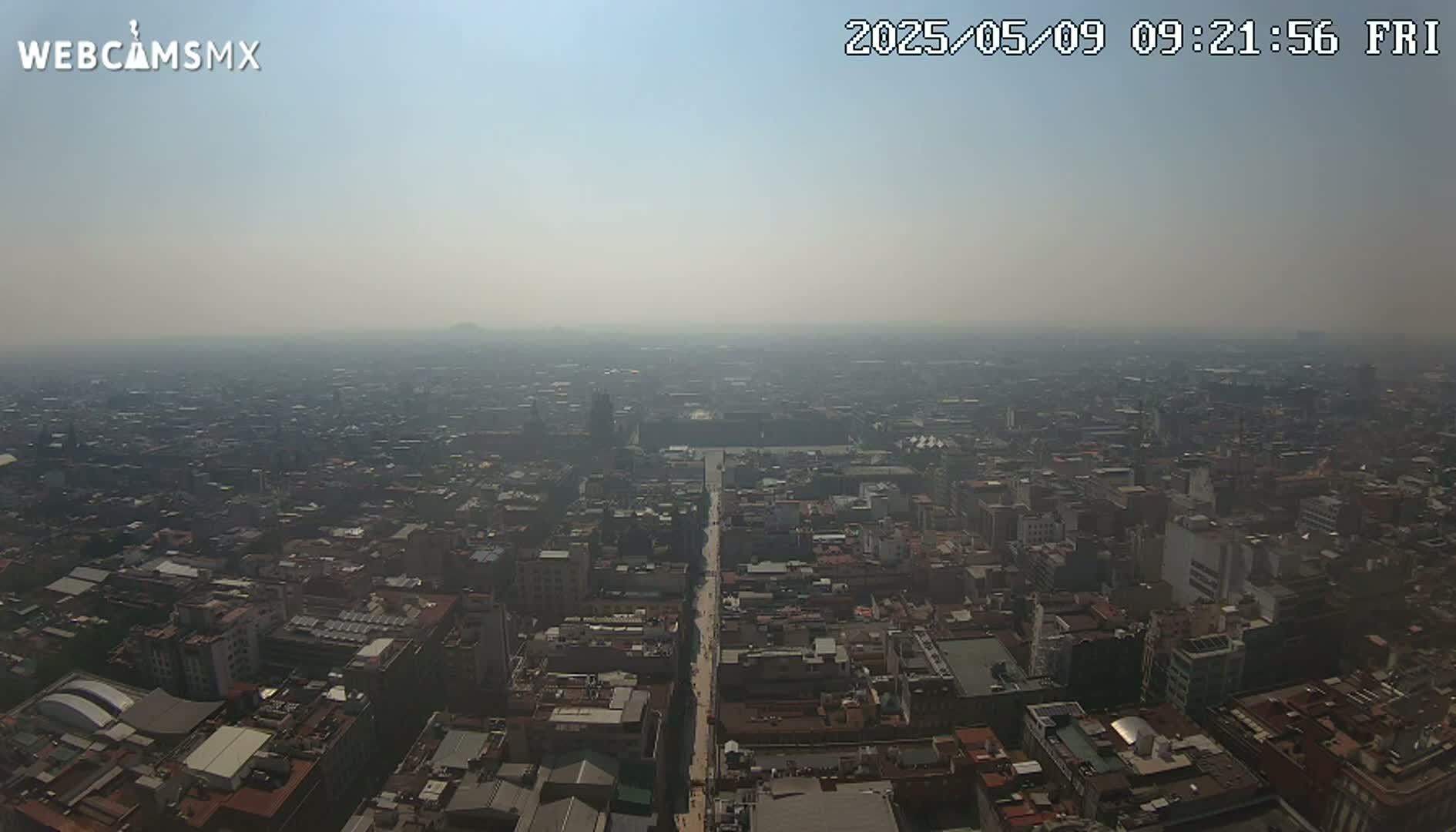 A hazy, high-angle view shows a sprawling city with many brown buildings and a long, straight road cutting through the center.