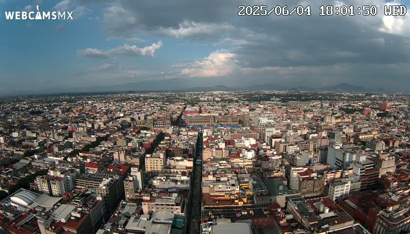 A sprawling city under a partly cloudy sky extends to distant mountains.