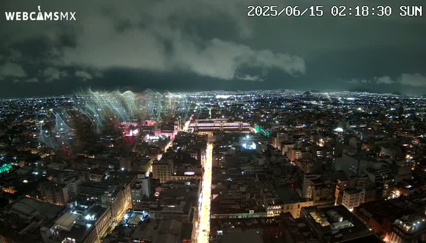 A nighttime, high-angle view of a sprawling city illuminated by lights, with a faint, ethereal glow in the sky above.