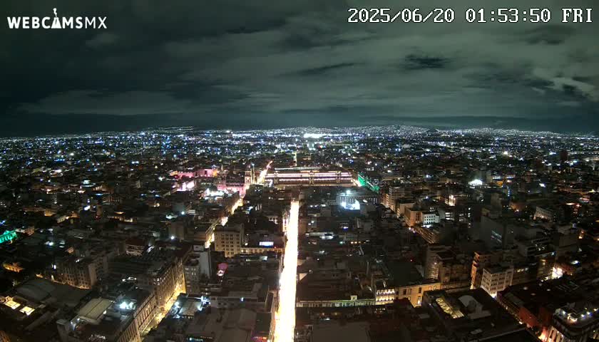 A nighttime aerial view of a sprawling city with a brightly lit street cutting through its center under a partly cloudy sky.
