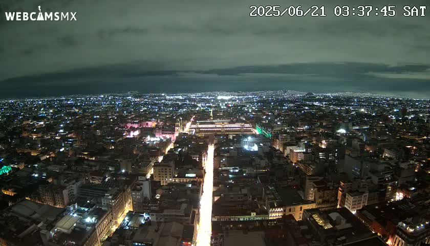 A nighttime aerial view of a sprawling city with a brightly lit street running through its center under a mostly cloudy sky.
