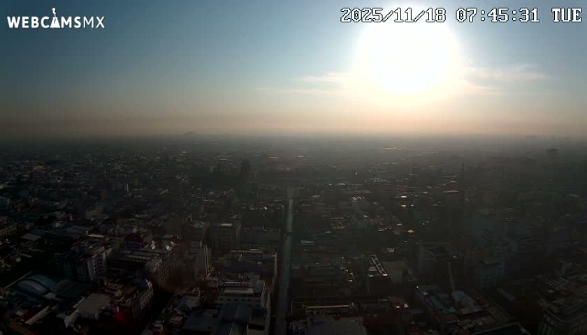 A high-angle, long shot shows a sprawling city under a cloudy sky with distant mountains visible on the horizon.