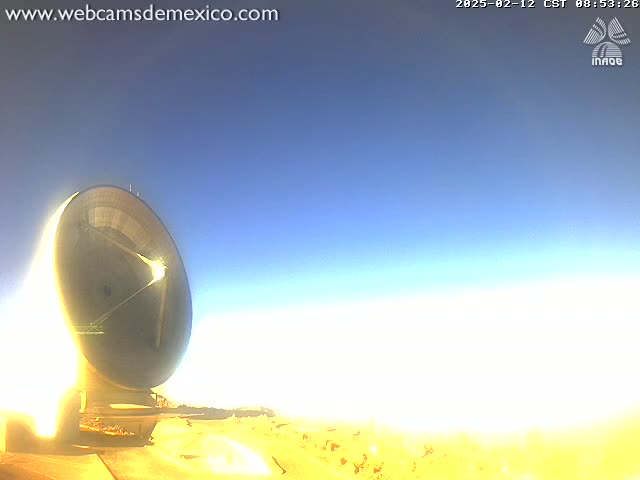 A large, dish-shaped telescope sits on a bright, sandy landscape under a clear, mostly blue sky.