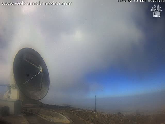A large satellite dish is visible in the foreground, partially obscured by fog under a partly cloudy sky.