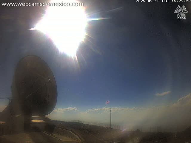 A large satellite dish is positioned in the foreground under a bright, sunny sky with some low-lying clouds and haze in the distance.