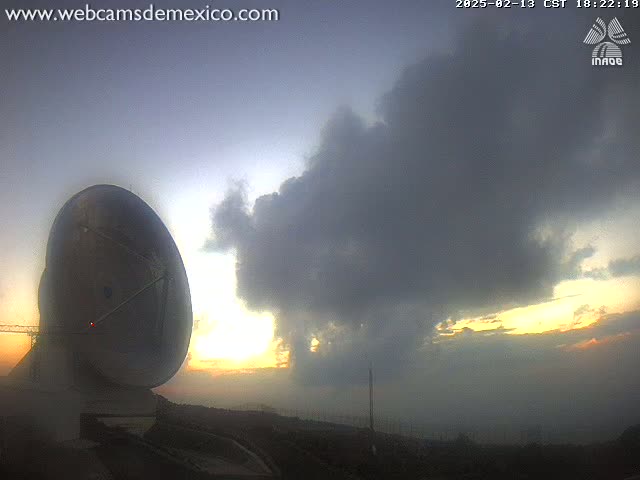 A large satellite dish is silhouetted against a hazy sunset with a large, dark cloud overhead.