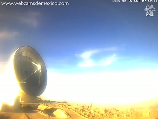 A large, dark-colored satellite dish sits on a mostly barren, light-brown landscape under a mostly clear, bright blue sky with a few wispy clouds.