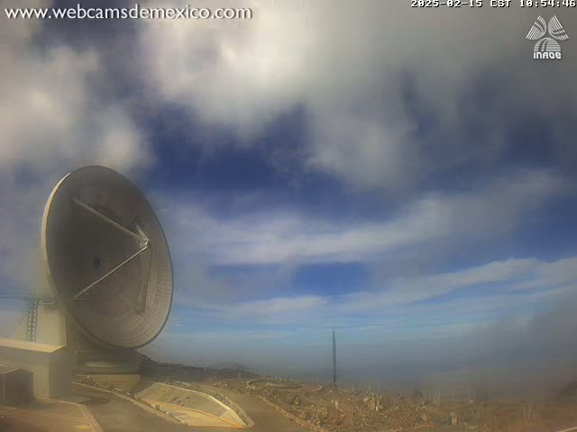 A large satellite dish sits on a hilltop under a partly cloudy sky with hazy conditions in the distance.