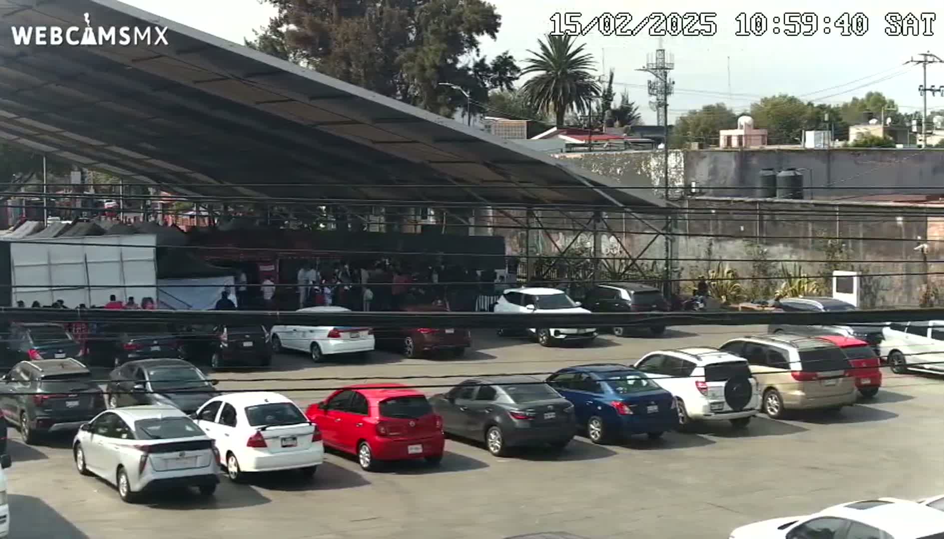 A partly sunny day reveals a large parking lot full of cars under a covered structure, with a crowd of people gathered near a building in the background.