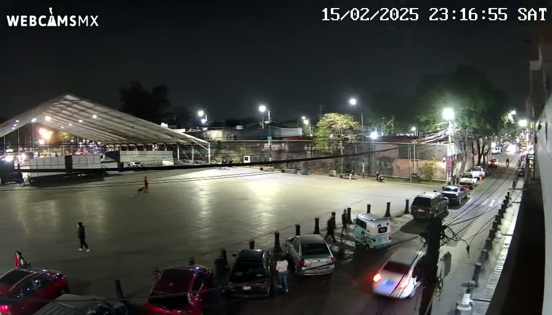 A nighttime view of a mostly empty plaza with cars parked along the sides, a large tent structure in the background, and a few pedestrians.