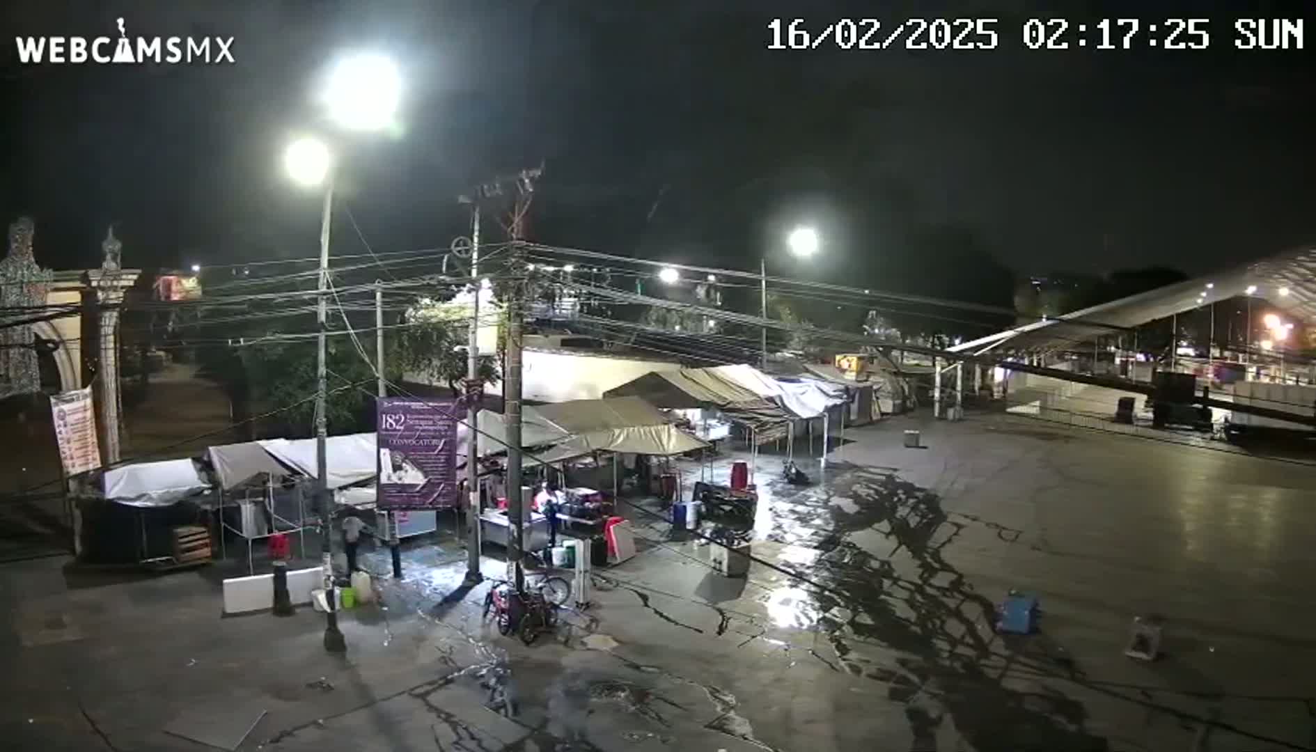 A wet nighttime scene shows several temporary market stalls set up in a plaza under bright streetlights.