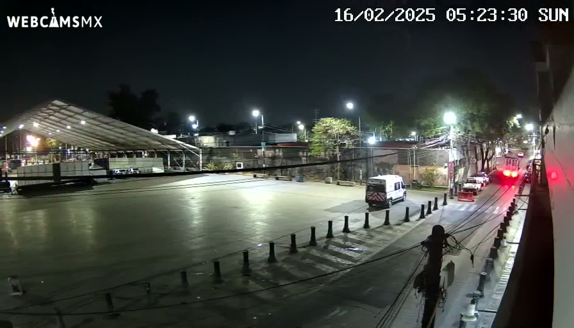 A nighttime, outdoor scene shows a large empty paved area next to a street with several vehicles, including a van, and a large covered structure in the background under artificial light.