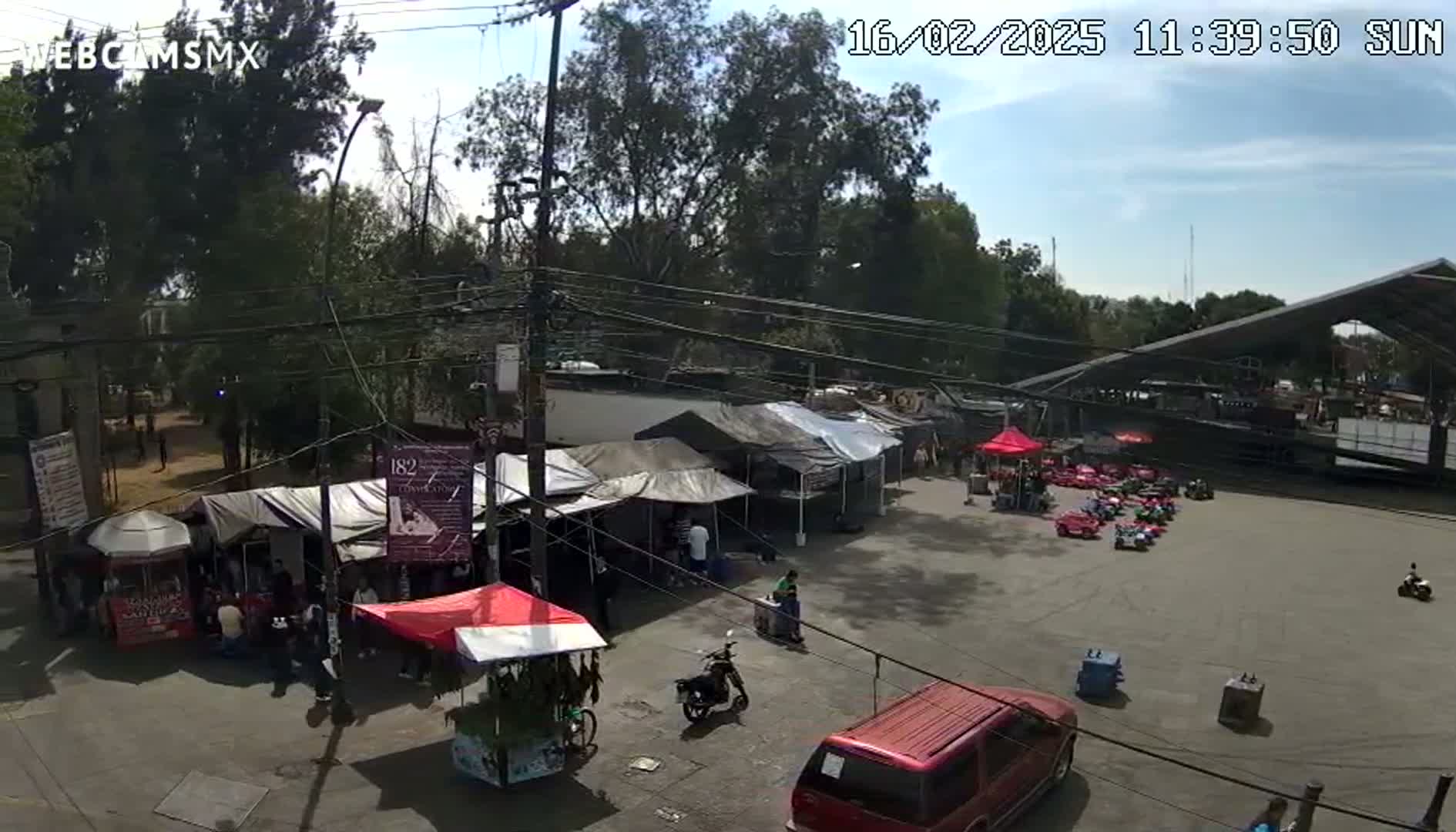An outdoor market scene under sunny skies features numerous vendor stalls, a few people milling about, and a line of small red cars parked in a nearby lot.