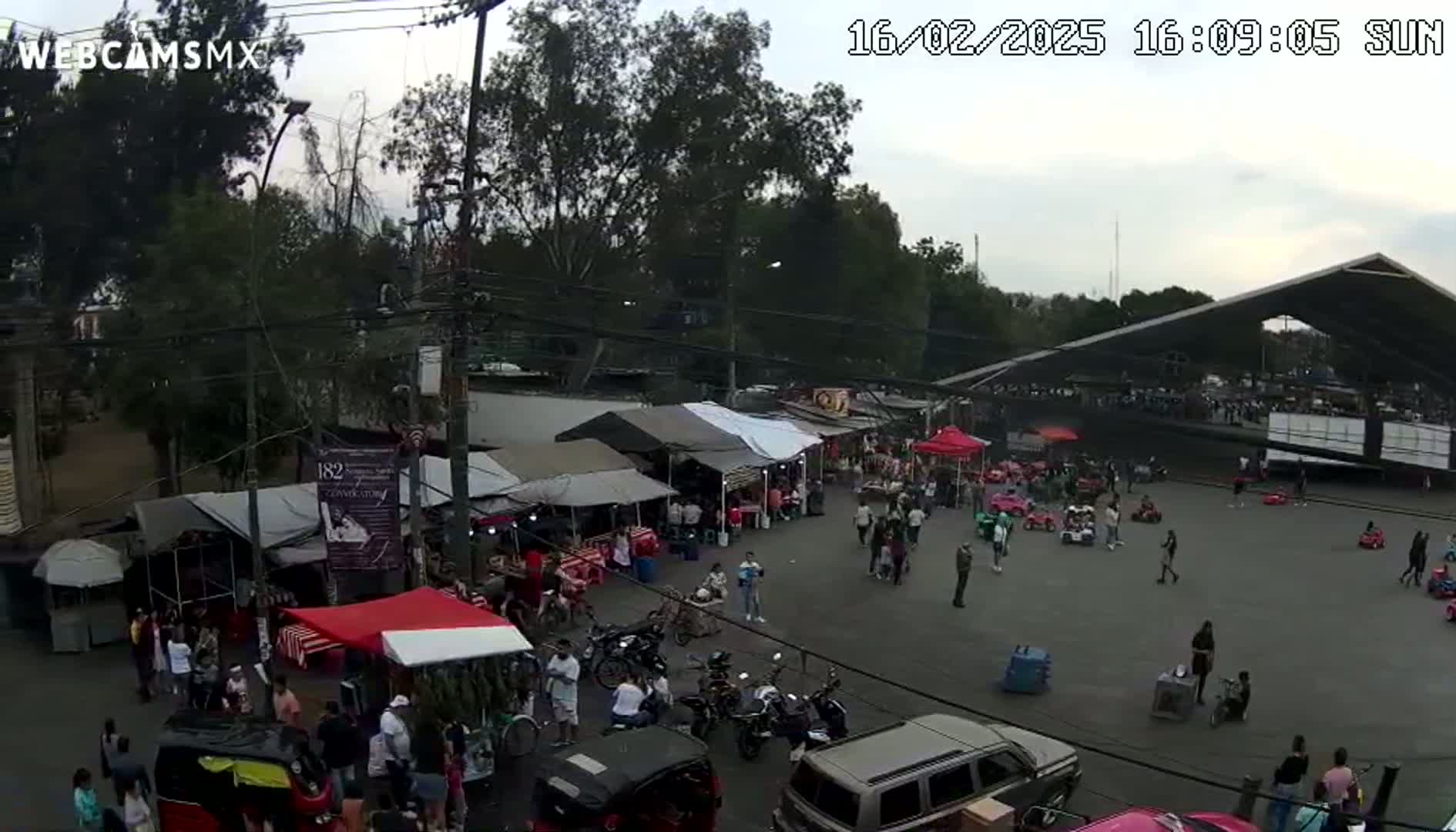 An outdoor market scene under an overcast sky, with numerous stalls, people browsing, and vehicles parked, near a large covered structure.