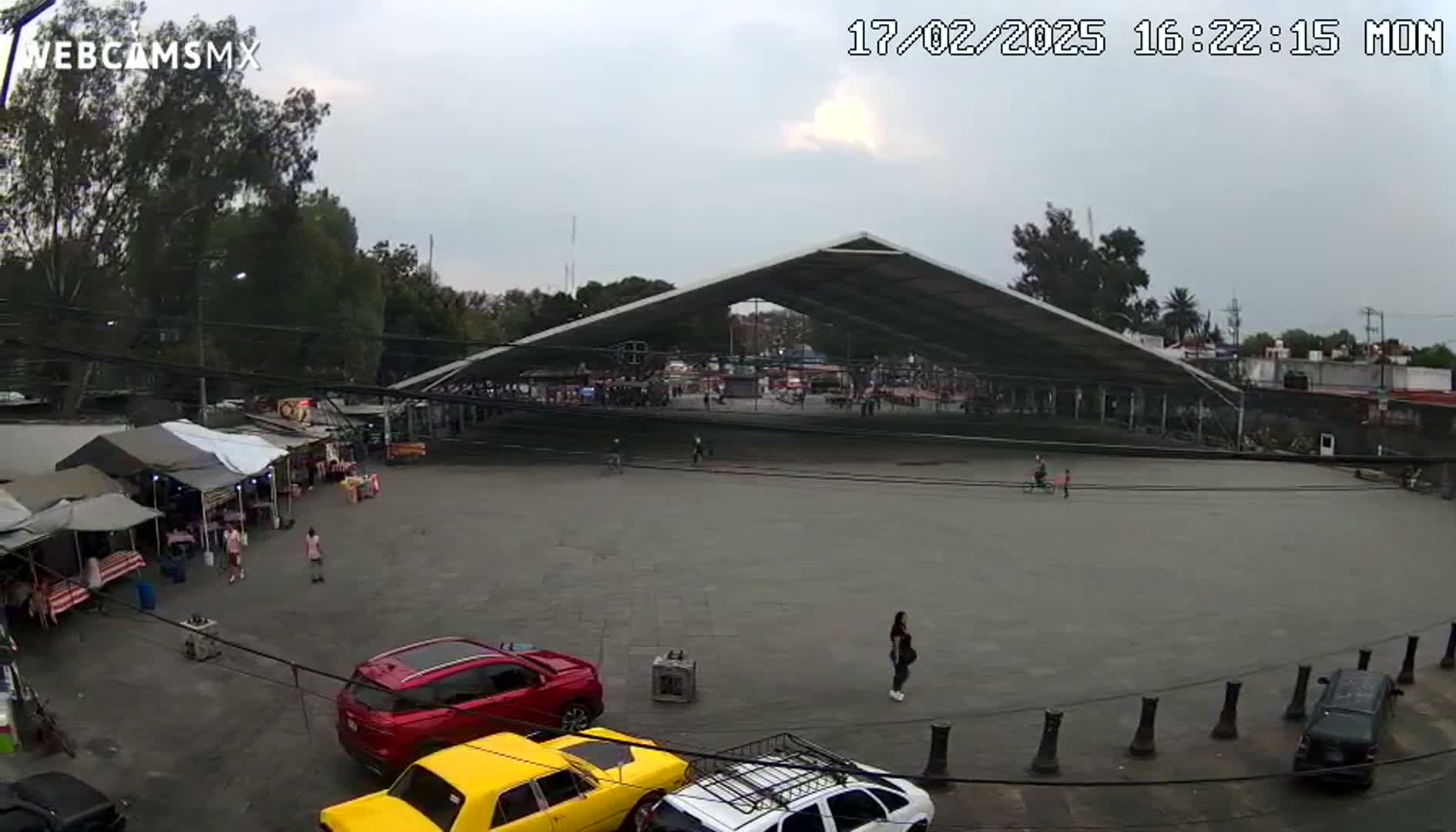 A large, open-air plaza with a covered structure in the center, surrounded by trees, market stalls, and parked cars, under an overcast sky.