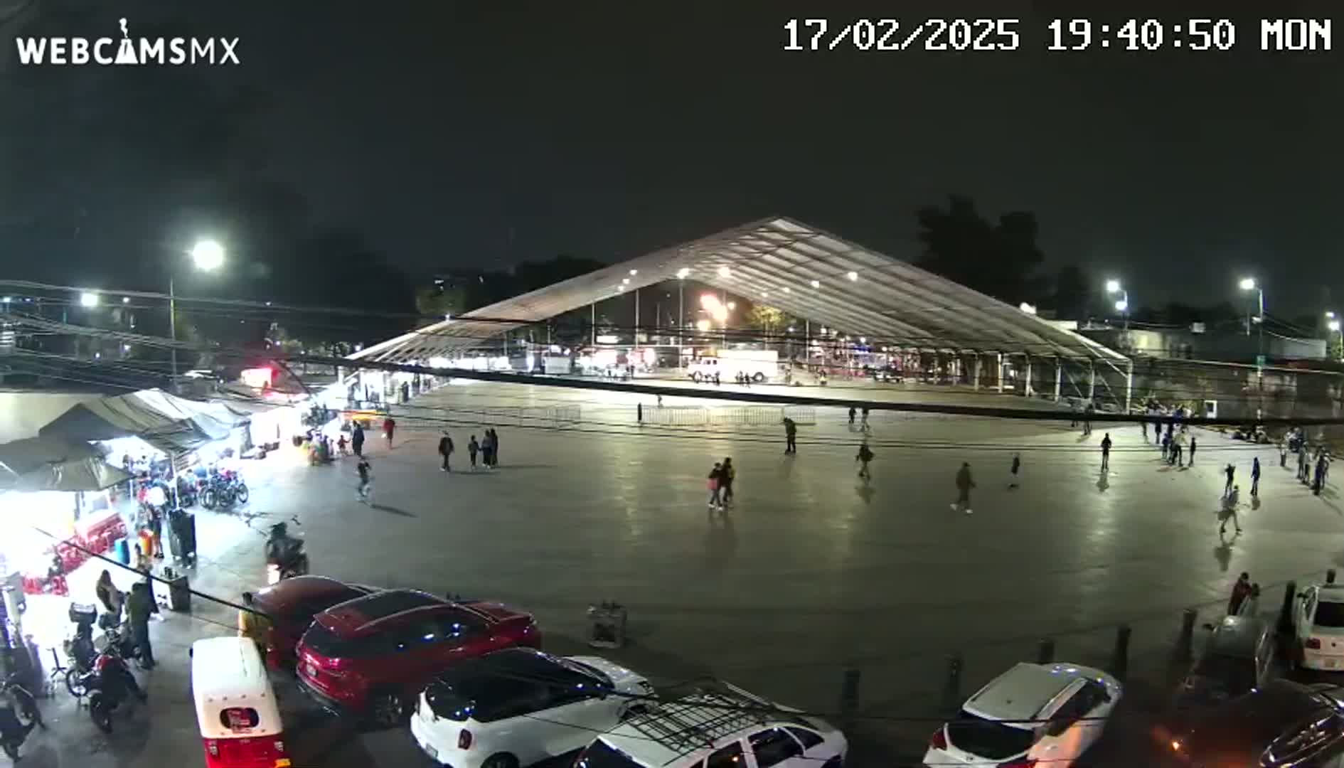 A nighttime aerial view shows a large, open paved area with a few people walking around, some parked cars, and a large tent structure, surrounded by smaller tents and food stalls.