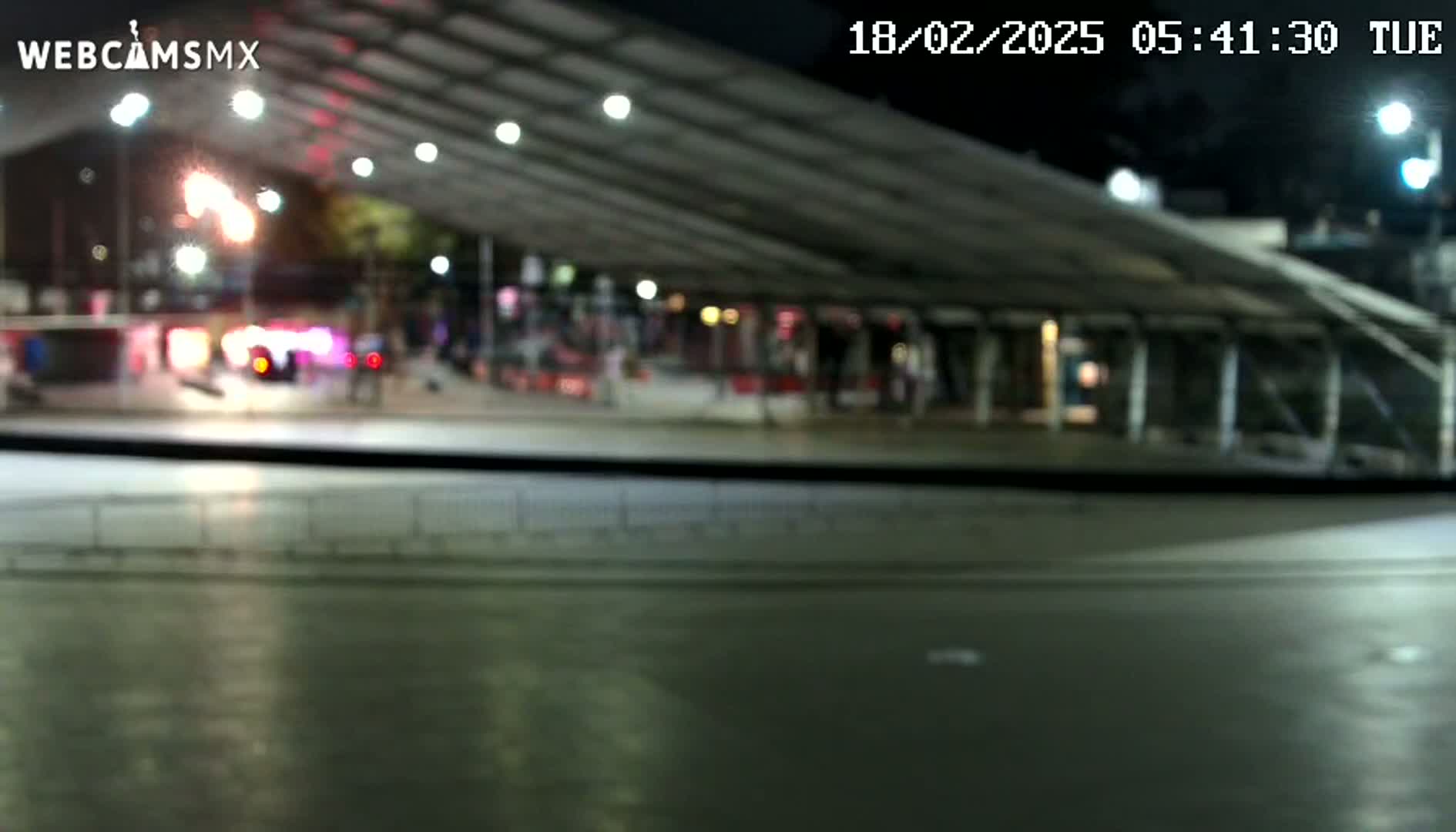 A blurry nighttime view from inside a vehicle shows a mostly empty plaza under a large covered structure, illuminated by artificial lights.