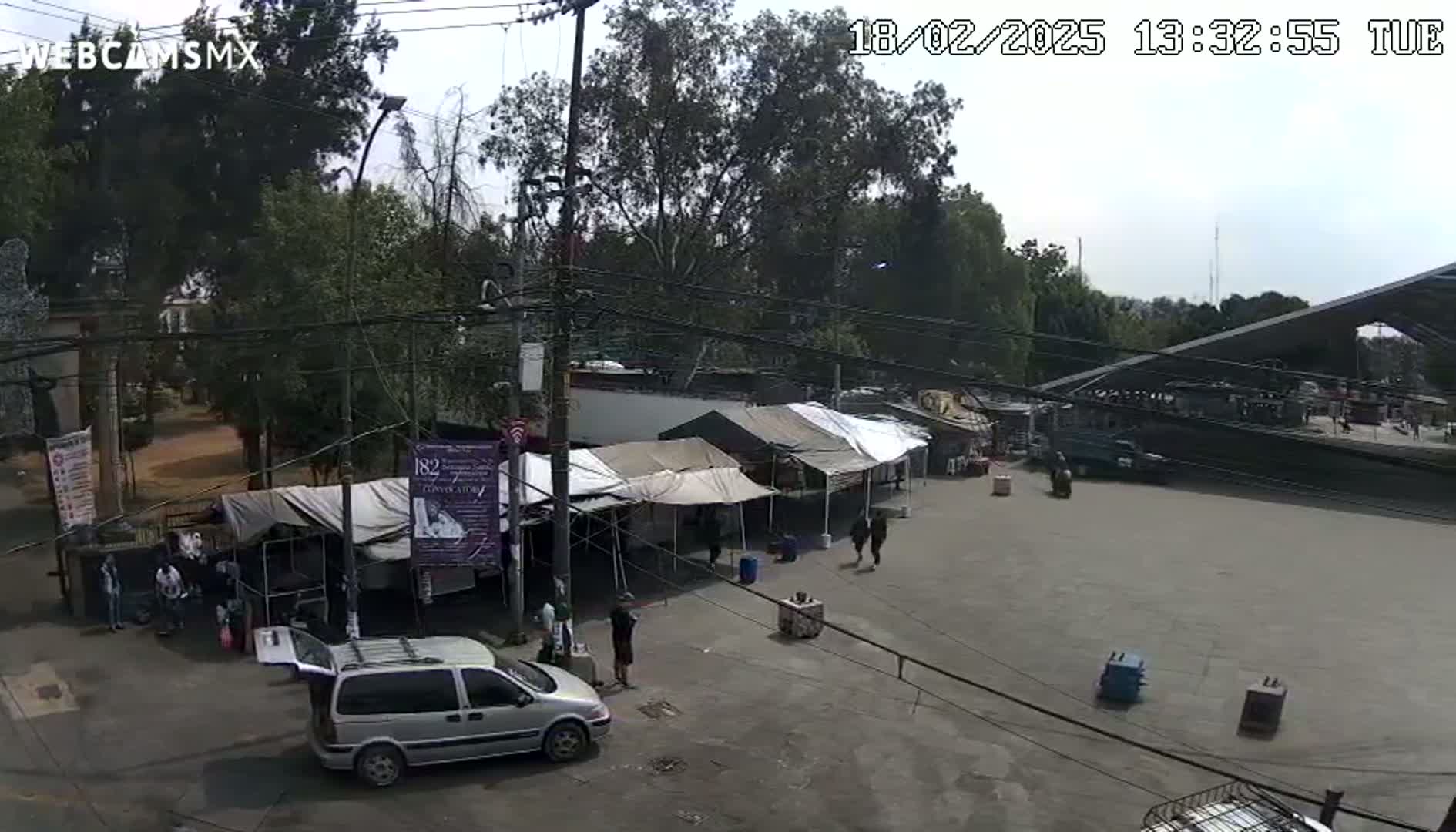 An outdoor market with several tents set up in a paved area under a partly cloudy sky, surrounded by trees and a minivan parked nearby.