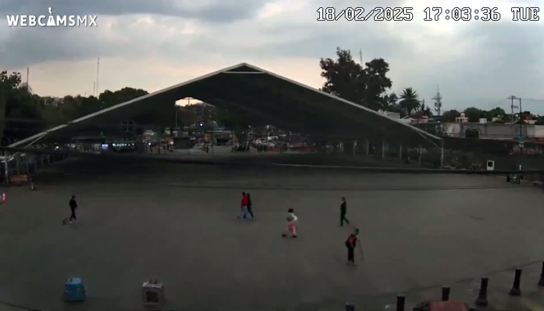 A large, open-air structure with several people walking across a mostly empty paved area under an overcast sky.