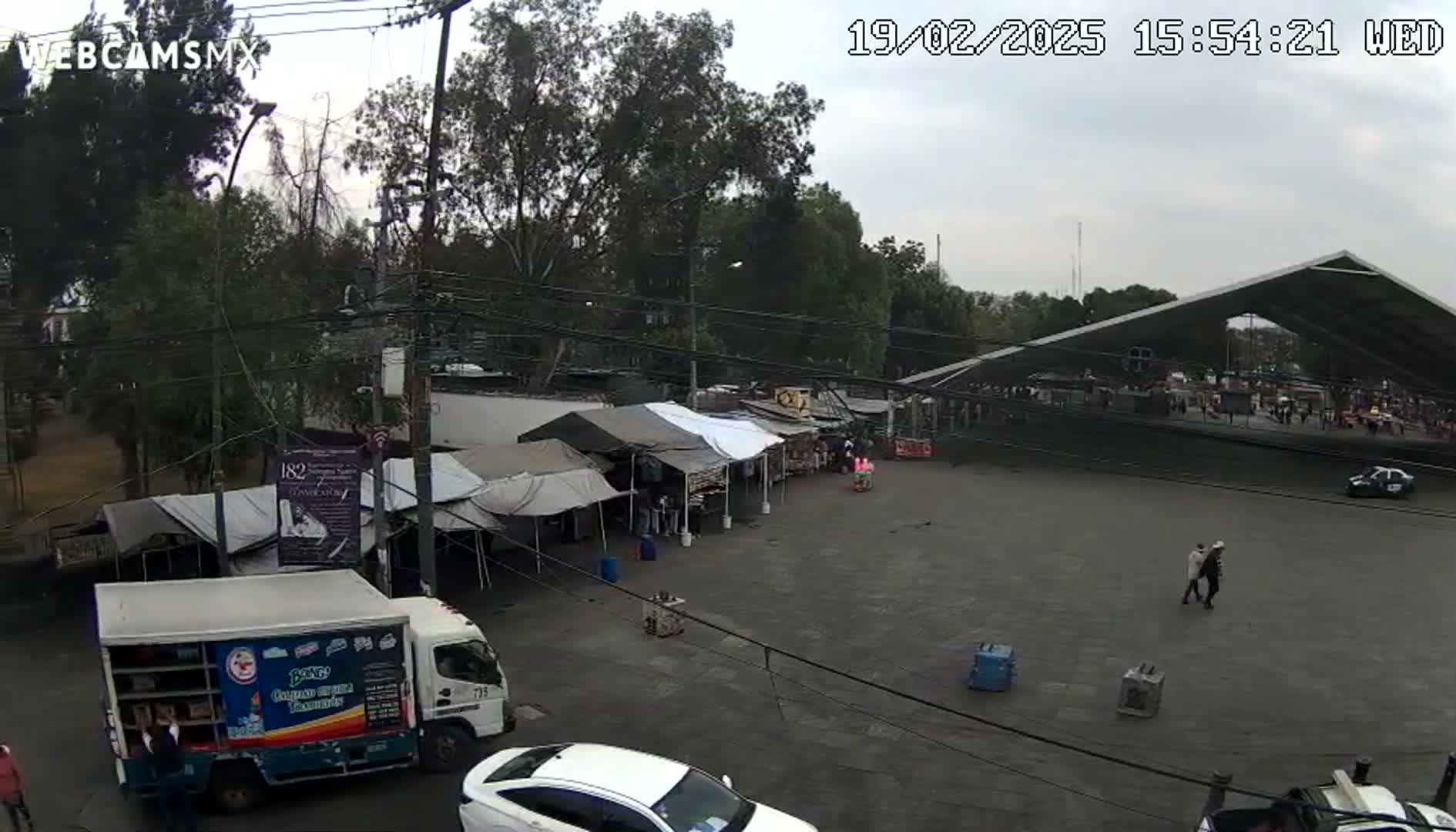 An overcast day reveals a street market scene with numerous tents, vehicles, and people, partially sheltered by a large, modern structure.