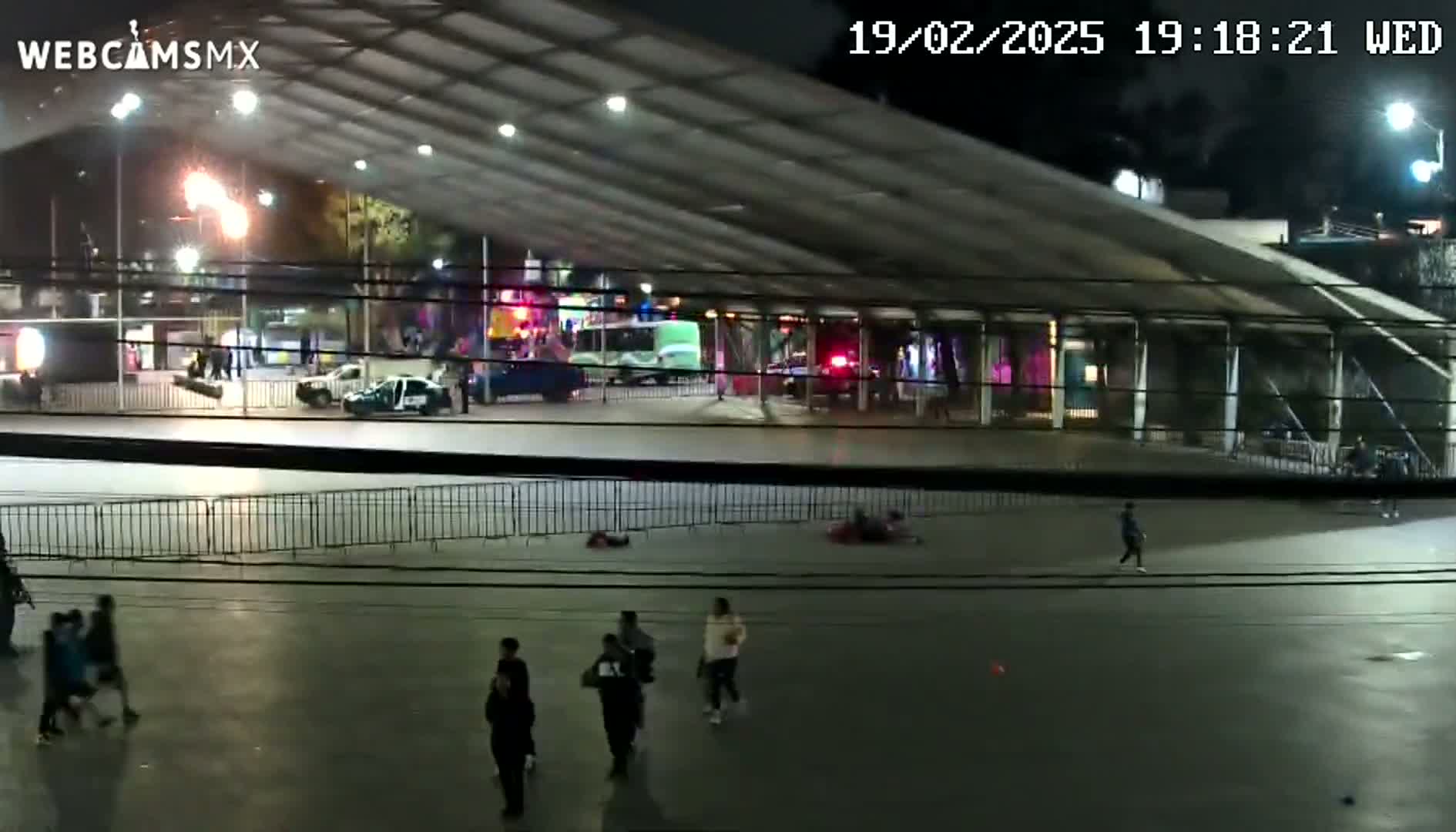 At night, under a large covered structure, several people are walking in a paved area near parked cars and emergency vehicles, with some people lying on the ground.
