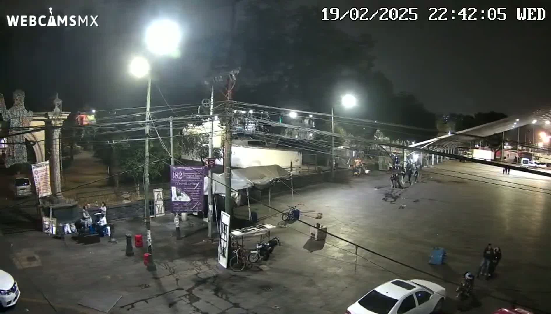 A nighttime, outdoor scene shows a mostly empty plaza with a few people gathered in small groups, vehicles parked along the edges, and various structures including a covered area and what appears to be a temporary market stall.