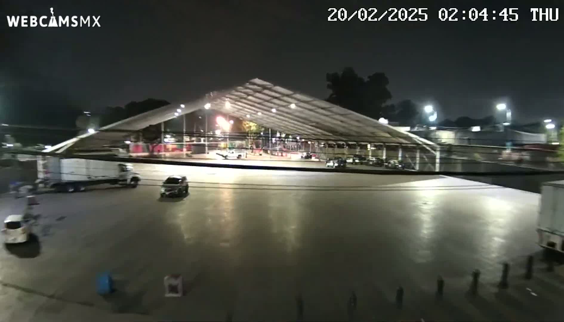 A nighttime view of a large, empty paved area under a large, illuminated tent-like structure with several vehicles parked nearby.