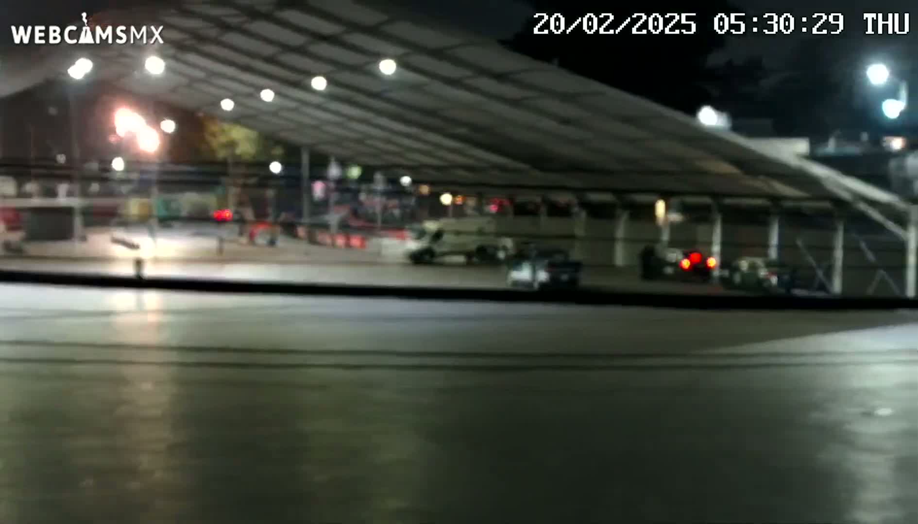 A nighttime view from inside a vehicle shows a mostly empty, covered outdoor area with several cars parked and a few people visible in the distance under artificial lighting.