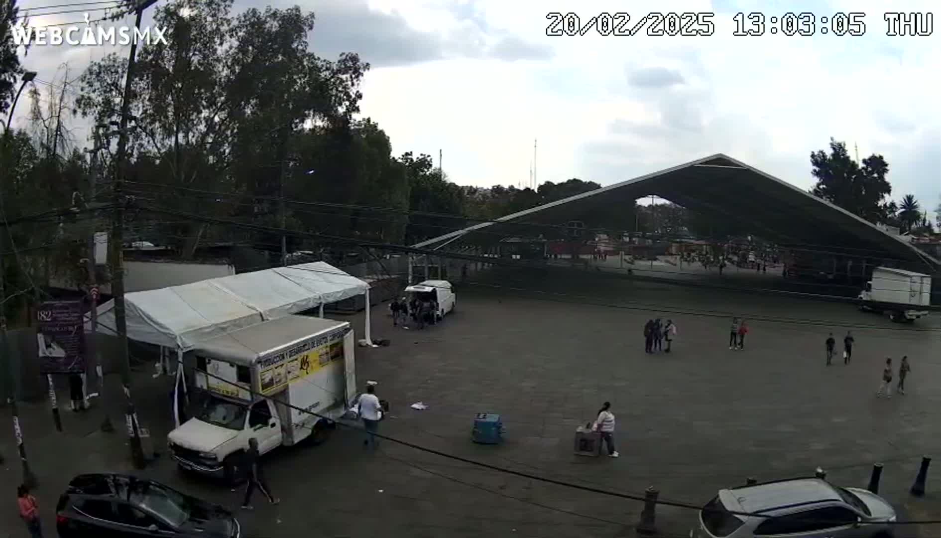 An overcast day shows a large paved area with several people, vehicles, and tents, under a large, modern roof structure surrounded by trees.