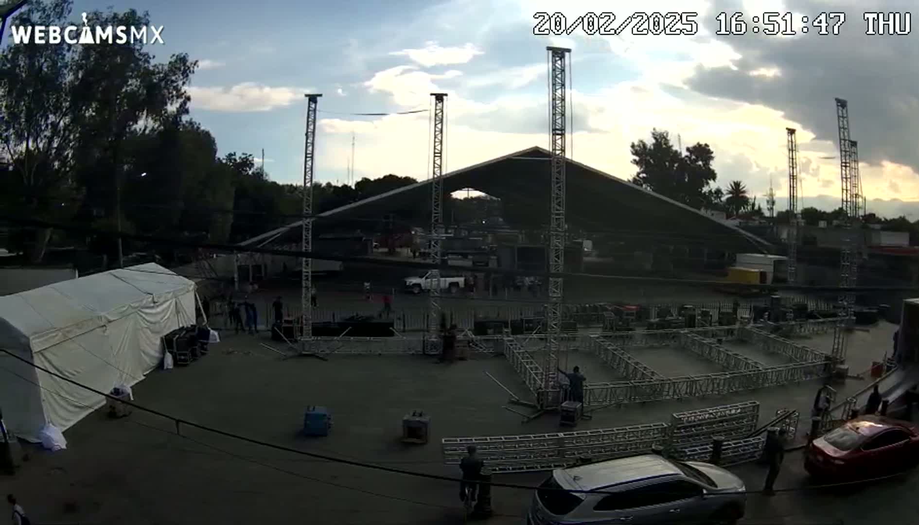 An outdoor area with partly cloudy skies is being set up for an event, featuring a large tent, multiple metal scaffolding structures, and several vehicles.