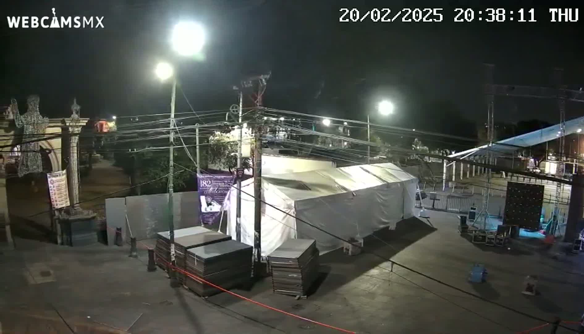 At night, under bright lights, a large white tent sits in a paved area near stacks of dark-colored materials, with some structures and streetlights in the background.