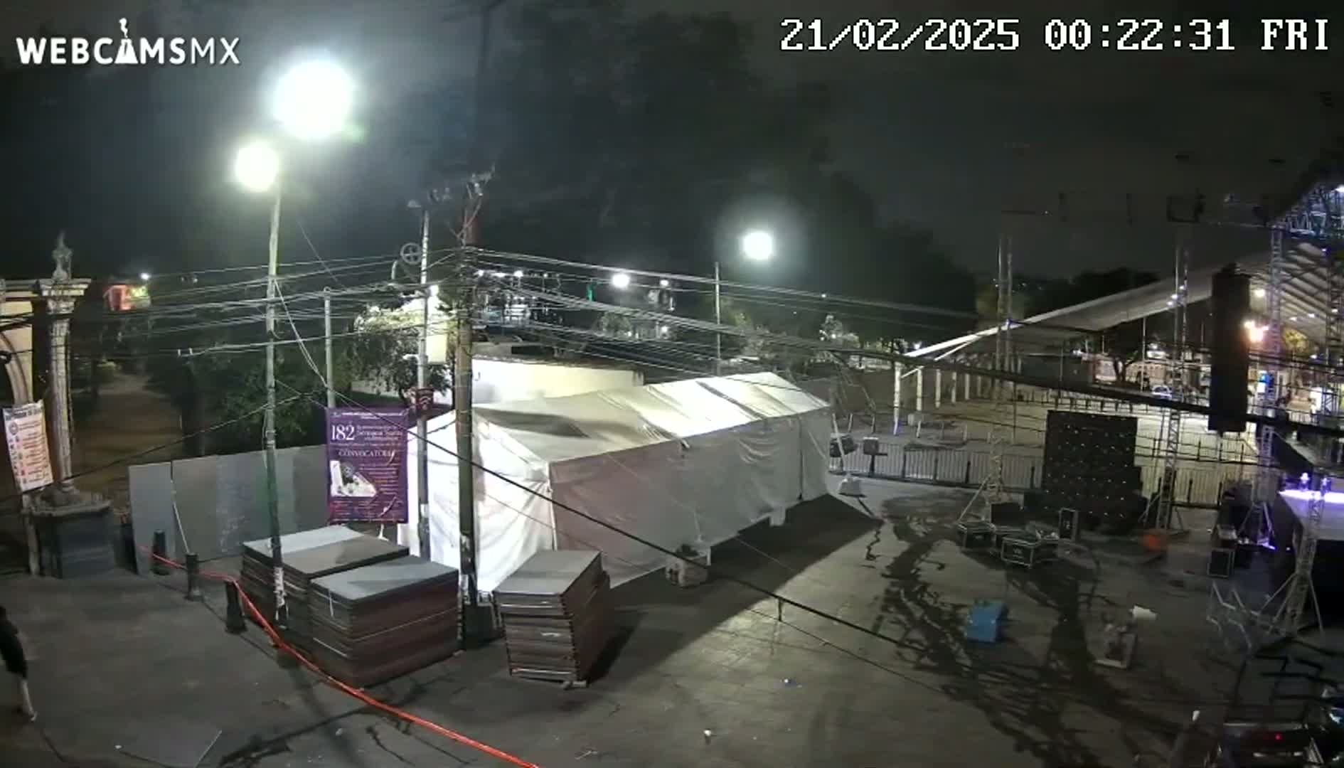 At night, under artificial light, a large white tent sits in a paved area near a stage being set up for an event, surrounded by various equipment and stacks of what appear to be flat panels.
