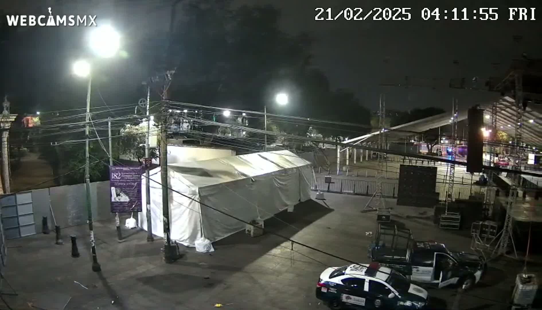 A large white tent sits in a paved area at night, under bright lights, near two police cars and various staging equipment.