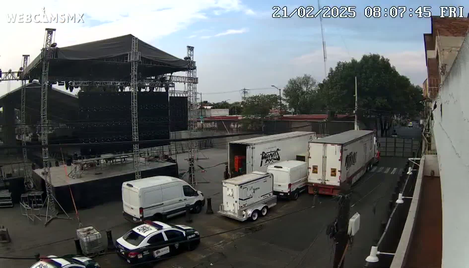 An outdoor scene under partly cloudy skies shows a large stage being set up, surrounded by several trucks, vans, and police cars.