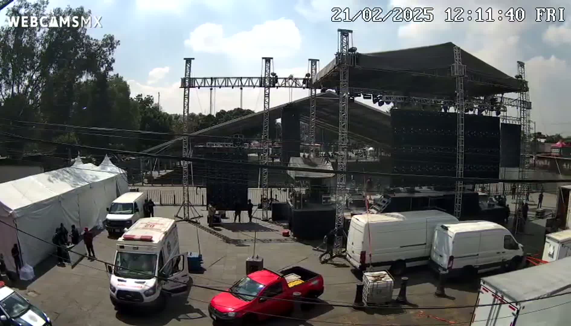 An outdoor scene under partly cloudy skies shows a large stage being set up, surrounded by various vehicles, including vans and an ambulance, near a large white tent.