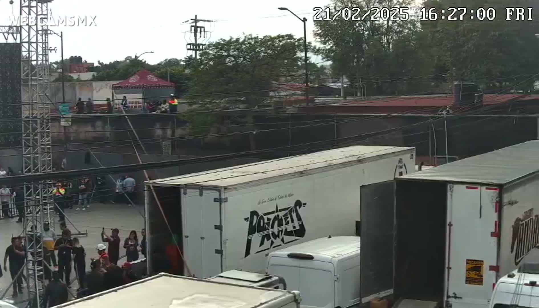 An overcast day shows several people gathered in an outdoor area near large trucks and staging equipment.